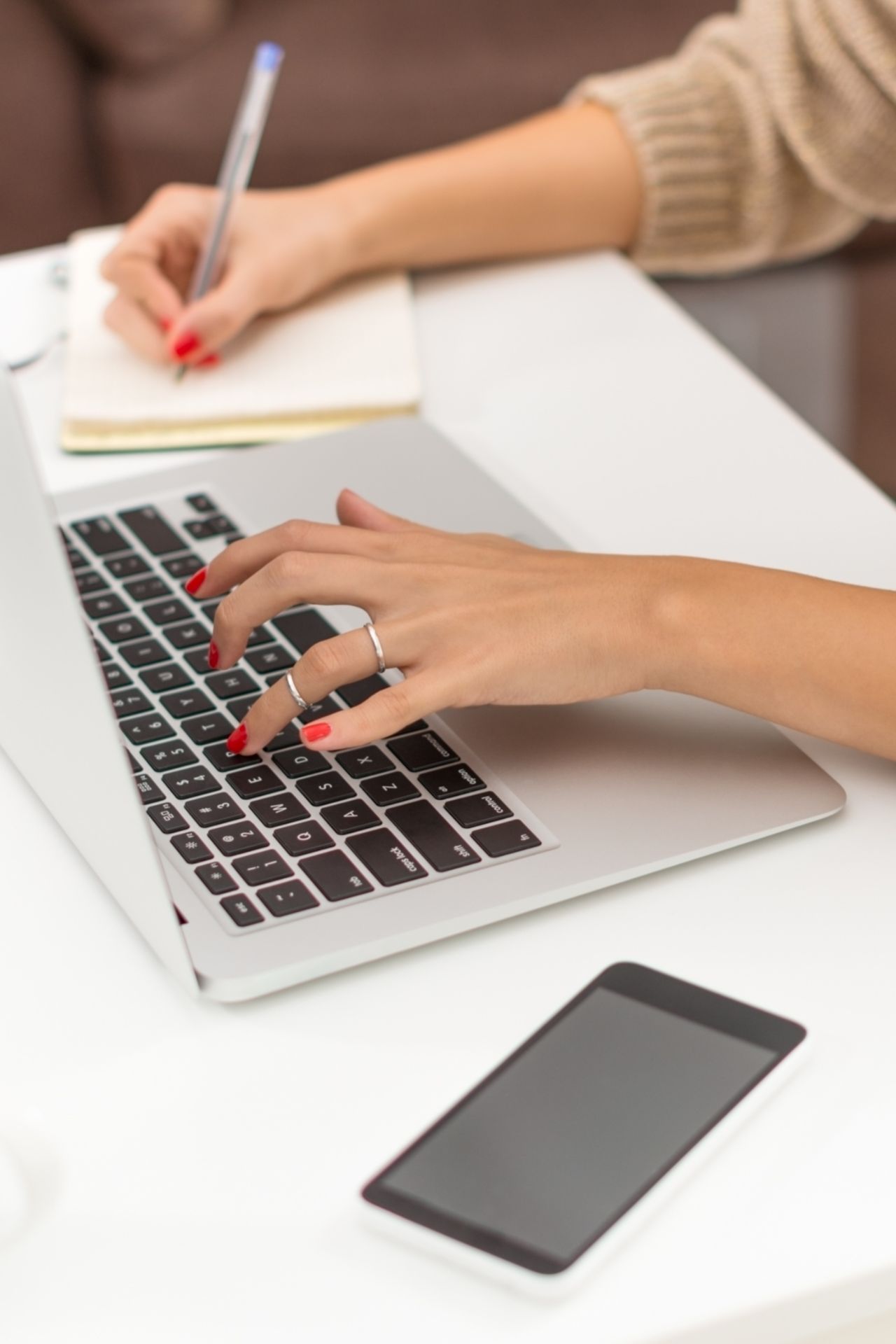 Női kéz piros köröm, laptomon gépel, másik kezével füzetbe ír - önismereti coaching online és Karcagon.