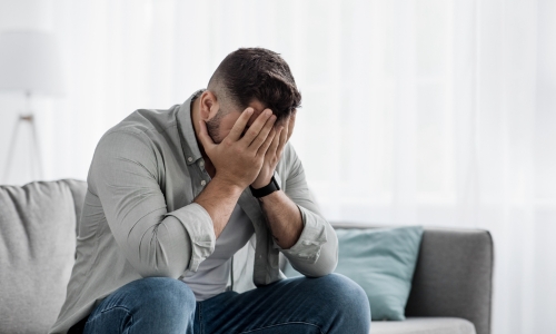 Férfi a kanapén ülve, arcát a tenyerébe rejti el, fél segítséget kérni - life coaching online és Karcagon.